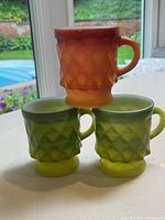 Photo showing two green and one orange FireKing collectible glass mugs with diamond texture pattern.