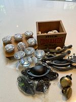Assortment of drawer knobs and pulls including porcelain, glass, and metal knobs and pulls, displayed on a white surface next to a wooden box.