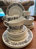 Full view of stacked plates, bowls, cups, creamer and gravy boat with black floral botanical pattern on white ceramic.