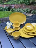 Full view of the mustard yellow Melmac dish set arranged on a black outdoor table in a garden setting, showing all included pieces.