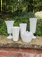 Front view of 5 white milk glass vases of assorted shapes and sizes displayed outdoors on stone ledge