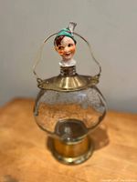 Front view of the musical liquor bottle showing the porcelain leprechaun head with green hat, crackled glass globe, and brass base and top with handle.
