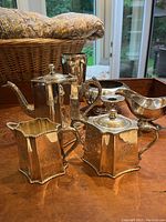 Front and side view of engraved silver-plate tea set with teapot, creamer, and sugar dish plus assorted silver items in background