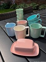Photo showing a group of pastel colored vintage Melamine dishes including mugs, bowls, and a butter dish displayed on outdoor table.