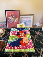 Overall display of the lot showing framed Suchard print with red background, framed beach print, canvas butterfly artwork on floor, Bud Man posters in front, and Charlie Brown poster partly visible.
