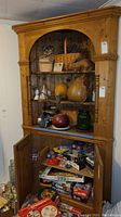 Full view of vintage wooden corner cabinet filled with decorative items on shelves and board games inside lower cabinet doors.