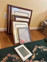 Assorted picture frames stacked against wall showing variety of sizes from small to large with wood-tone and white frames.
