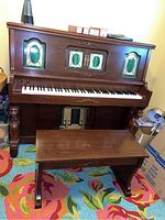 Front view of player piano showing carved legs, decorative green panels, and matching wooden bench on a colorful rug.