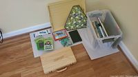Photo of all items including books in plastic bin, educational tablets, triangular floral box, butterfly puzzle tray, black journal, whiteout, and wooden art supply case on floor