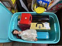 Top view of multiple carrying cases and a vintage dressed doll in white gown inside a plastic bin