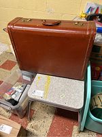 Photo showing the vintage brown suitcase standing upright on the children's table and nearby floor surface, visualizing the suitcase size and condition with visible scuffs and scratches.