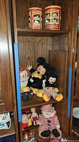 Wide shot of wooden shelf displaying vintage plush toys (Mickey Mouse large and small, Raggedy Ann, clown), small figurines, and collectible tins.