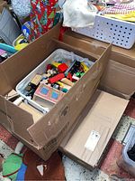Cardboard box containing white cloth basket filled with assorted wooden toy train pieces, vehicles, and houses.