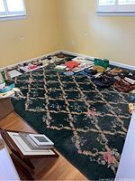 Photo of the full green wool area rug with a floral lattice design placed on a hardwood floor in a room with pale yellow walls, various items around the edges for scale.