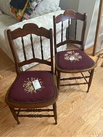 Two antique wooden chairs with needlepoint seats shown side by side on wood floor near cream couch. Both have burgundy seats with floral embroidery. Wood is medium brown with spindle backs and turned legs.