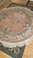 Round pink wool rug with floral and decorative border design, cotton fringe visible, placed on larger rectangular multicolor rug