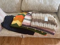 Four blankets folded and stacked on a beige couch, showing a colorful chevron, a striped and a crochet blanket and a solid black one underneath.