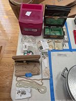 View of various costume jewelry items including faux pearl strands, earrings, watches, snake pin, replica coins, wooden jewelry box, plastic drawers, and a clear glass dish on a table.