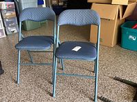 Two blue metal frame folding chairs with fabric seats and backrests, standing side by side on a carpeted floor in a storage area with boxes around.