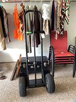 Front view of the black hand truck standing vertically with large chunky tires and vertical handle bars, surrounded by various household items.