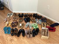 Full assortment of 24 pairs of women's shoes of various styles and colors arranged on wooden floor including sandals, flats, slip-ons, some with tags, some worn