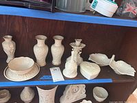 Full view of nine white Lenox porcelain items including vases, bell, bowl, dishes, and candlestick on wooden shelf.