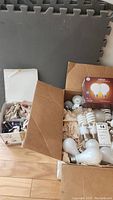 Open box containing various light bulbs including candelabra bulbs, HD light bulbs, and loose bulbs alongside four grey foam floor mats.