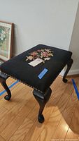 Black wooden bench with cabriole legs and needlepoint upholstery with floral motif, nail head trim visible on side edges.