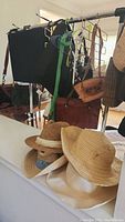 Photo showing four different straw hats with various trims and linings on a white surface with purses hanging on a rolling rack behind them.