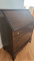 Side angled view of vintage wooden 3-drawer secretary desk showing hinged flip-top writing surface closed and overall condition.