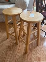 Two wooden stools standing side by side on hardwood floor near couch, showing front views and leg structure.
