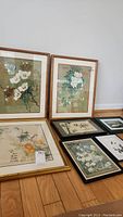 Photo displaying seven framed floral and Asian influence art pieces of varying sizes arranged on wooden floor.