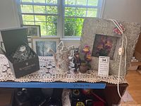 Entire lot viewed on top of white crocheted doilies on a shelf near window. Includes the clock box, resin monkey figurine, ceramic watering can, lighted flower frame with attached cord and glass stirrer rod.