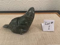 Front side view of the dark green soapstone walrus sculpture on a beige fabric surface, showing detailed carving and texture on the face and body, next to a small sign labeled 'LOT # 1'.