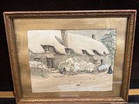 Framed watercolour artwork showing a snow-covered cottage in a winter setting with a figure near the door.