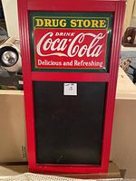 Front view of Coca Cola Drug Store chalkboard with red frame and metal Coca Cola sign at top, black chalkboard below.