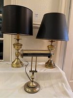 Three brass lamps: Two tall table lamps with black shades and one shorter bankers lamp with horizontal shade in brass and black.