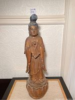 Full view of wood Asian lady carving standing on lotus base against wall with textured wallpaper background.