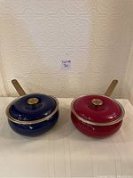 Two enamel-coated Copco pots with lids shown from front, colors navy blue and maroon with brass-tone handles.