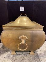 Front view of large vintage brass container showing dome lid and decorative ring handle with shell motif.