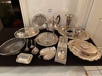 Wide angled photo showing assortment of silver plated serving trays, teapot, bowls, utensils, and decorative pieces arranged on a black surface against a textured wall.