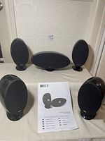 Four KEF HTS3001 satellite speakers and one KEF HTC3001 center speaker arranged with user manual, all with speaker grille covers attached.