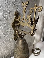 Full view of vintage brass bell mounted on ornate bracket with angel figure and chain pull against textured wallpaper.