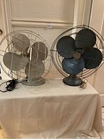 Two vintage 4-blade metal fans on a white cloth, one dark gray and one light gray, both showing wear.