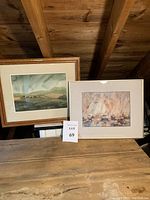 Two framed watercolor artworks displayed side-by-side on a wooden surface under a slanted wood ceiling, one landscape and one seascape with sailboats.