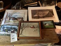 Five framed artworks arranged on a wooden surface under attic roof beams, showing various farm and barn scenes in winter and summer.