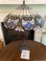 Full view of lamp on table showing stained glass shade with blue, gray, pink, and white glass in heart patterns and dark ornate metal base.