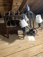 Wooden mid-century magazine rack next to a black bronzed metal chandelier with three white shades lying on wooden floor.