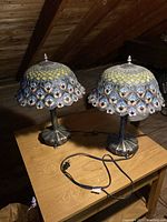 Two Tiffany style peacock feather table lamps on wooden table under angled lighting