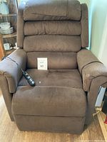 Full view of brown recliner chair showing plush segmented backrest, cushioned armrests, and seat with upholstery showing minor wear and stains. Remote control resting on seat connected to chair by cable.
